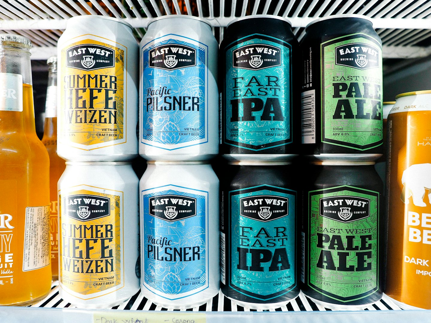 Reihe von Craft-Bierdosen im Kühlschrank mit verschiedenen Sorten von East West Brewing Company, darunter Summer Hefe Weizen, Pacific Pilsner, Far East IPA und Pale Ale.