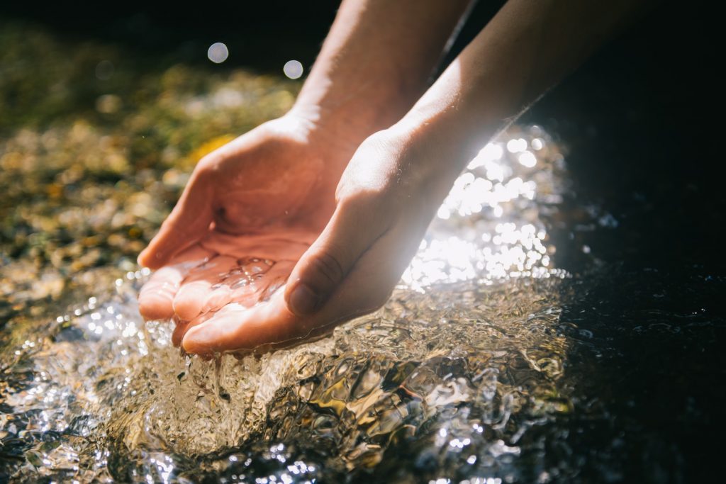 Hände, die sanft Wasser aus einem klaren Flussbett halten, mit Sonnenlicht, das auf die Wasseroberfläche reflektiert.
